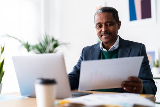 Concentrated Ethiopian male entrepreneur with laptop looking at document and analyzing data while sitting at desk and working on project in spacious workplace