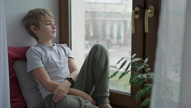 Teenager sitting next to window alone and is thinking about something at home. Sad boy is looking out of the window. Emotional concept