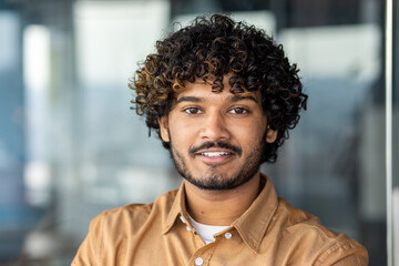 Young smiling indian programmer close up smiling and looking at camera, portrait of man with curly hair and shirt inside office at work, businessman entrepreneur with beard working on project