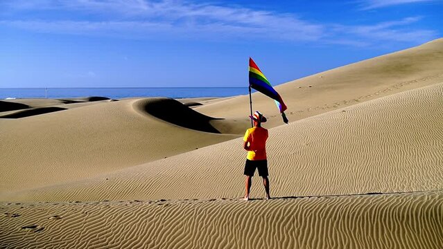 Gay boy proudly displaying rainbow flag outdoors in beautiful setting with sand dunes, ocean, seagulls and blue sky of Maspalomas, Canary Islands