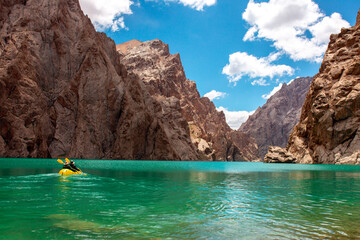 Obraz premium Kayaking on a mountain lake. Two men are sailing on a red canoe along the lake along the rocks. The theme of water sports and summer holidays.