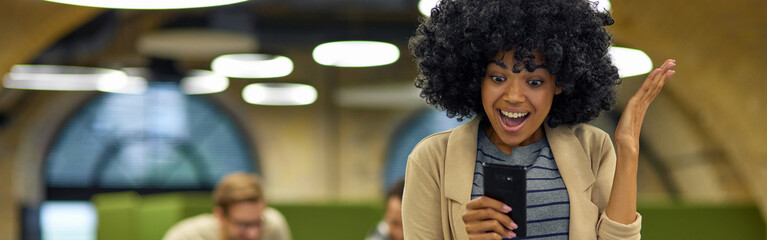 Good news. Young excited mixed race woman using smartphone and smiling while standing in the coworking space with her colleagues working on the background