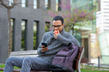 Fototapeta premium Young black man eating an apple and using mobile phone on bench outside