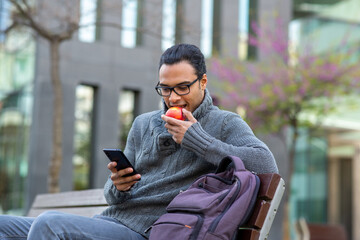 Young african man eating an apple and using cell phone while sitting on outside bench