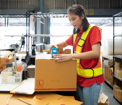 Warehouse staff packing goods for shipping order dispatch. Asian woman working in storehouse sealing cardboard box for shipment delivery. Logistics and supply chain distribution management background.
