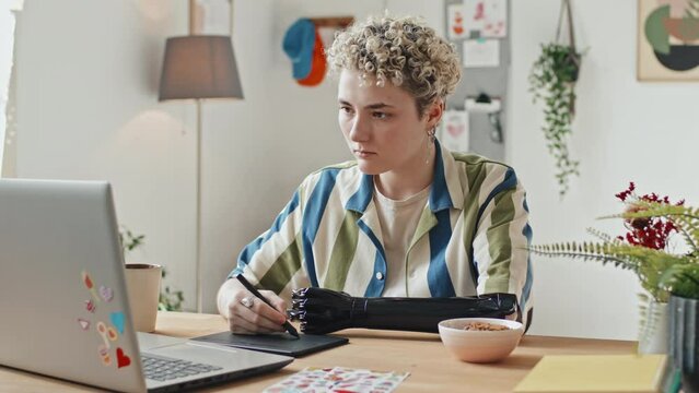 Portrait of modern gen Z girl with disability sitting at desk in front of laptop drawing picture using graphics tablet