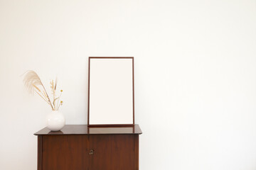 Minimalistic and stylish mock up poster frame standing on cabinet, concept with retro furnitures,brown cabinet,White walls, home decor. Nice interior of living room. Real photo. minimalism copy space