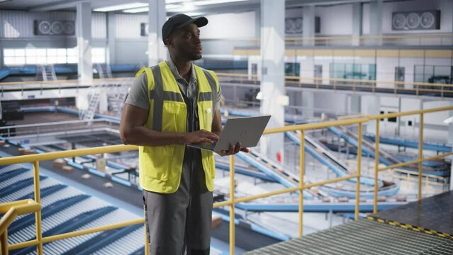African American Man Working in a Modern Spacious Logistics Center with Automated Belt Conveyors for Sorting Orders. Black Male in High Visibility Vest Using Laptop Computer in an Industrial Facility