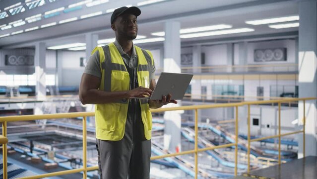 Young Black Man Standing on a Metal Platform, Using a Laptop Computer at a Post Office Logistics Center. African Specialist Working at a Sorting Facility, Configuring the Automation Process