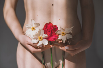 A woman holds daffodils and a red tulip in her hands close-up. The girl's hands cover the intimate areas with flowers - a symbol of innocence. Symbolic image of menstruation - obrazy, fototapety, plakaty