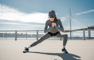 Obraz premium Stretching of legs and body training in sneakers, exercises of a woman in sportswear, cardio training. Against the background of a bridge in the city.