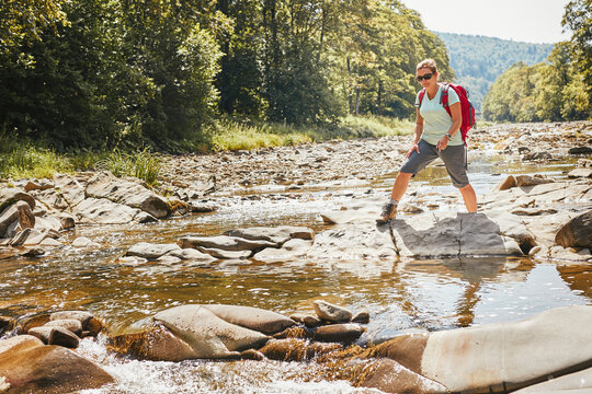 Trekking with backpack concept image. Female backpacker wearing trekking boots crossing mountain river. Woman hiking in mountains during summer trip