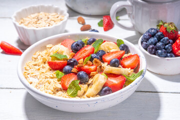 Bowl of sweet breakfast oatmeal with berry. Portion plate of wholegrain oats porridge, with yogurt, fresh banana, strawberry, blueberry, almond nuts, on white wooden table. Healthy summer morning food