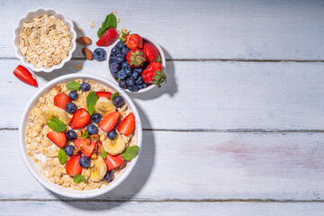 Bowl of sweet breakfast oatmeal with berry. Portion plate of wholegrain oats porridge, with yogurt, fresh banana, strawberry, blueberry, almond nuts, on white wooden table. Healthy summer morning food