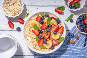 Bowl of sweet breakfast oatmeal with berry. Portion plate of wholegrain oats porridge, with yogurt, fresh banana, strawberry, blueberry, almond nuts, on white wooden table. Healthy summer morning food