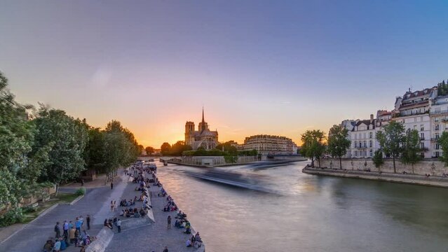 Rear view of Notre Dame De Paris cathedral at sunset with sun in the frame timelapse. Panoramic view from Tournelle bridge. People sitting on waterfront. Orange sky in background. Paris, France Europe