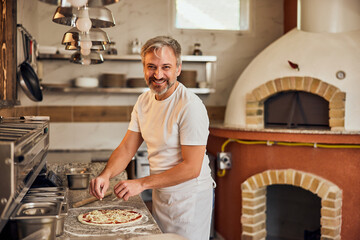 Photo of a smiling pizza master, working at the kitchen, making pizzas.