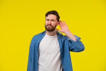 Young, serious, blond man in a blue shirt on a yellow background, holding an ear to listen better,...