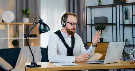 Attractive smiling experienced bearded married office worker in glasses sitting in front of computer and has video conference with friend or coworkers using headset