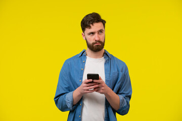 Bearded, cheerful, attractive guy in casual outfit, jeans shirt, holding smart phone in hands, using 3G internet, wi-fi, checking email, doing online shopping over yellow background