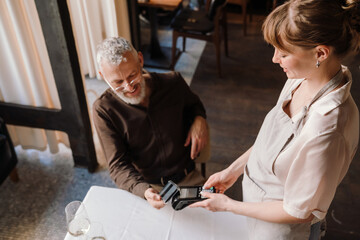 Obraz premium Cheerful young waitress accepts payment through bank terminal for lunch of happy mature man in cafe