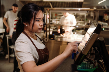 Cute asian business owner in apron punches food order on monitor in cafe