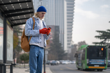 Caucasian millennial guy with backpack standing at bus stop using smartphone outdoors, checking public transport schedule in mobile app. City transportation and mobile devices concept