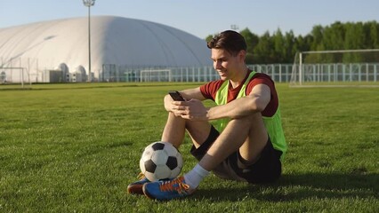 Portrait of young man football player sitting on soccer field and using smartphone