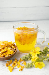 Herbal medicinal tea drink made of Verbascum thapsus, the great mullein, greater mullein or common mullein. Yellow dried flower petals. Glass cup with tea on white background and dried petals in bowl.