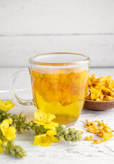 Herbal medicinal tea drink made of Verbascum thapsus, the great mullein, greater mullein or common mullein. Yellow dried flower petals. Glass cup with tea on white background and dried petals in bowl.