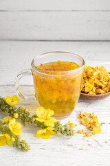 Herbal medicinal tea drink made of Verbascum thapsus, the great mullein, greater mullein or common mullein. Yellow dried flower petals. Glass cup with tea on white background and dried petals in bowl.