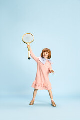 Full-length portrait of playful, happy, smiling little girl in pink dress posing with tennis racket over blue studio background. Concept of childhood, emotions, fun, fashion, active lifestyle, sport