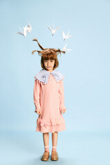 Full-length portrait of cute, lovely little girl, child in pink dress posing with paper birds above head against blue background. Concept of childhood, emotions, fun, fashion, lifestyle, imagination