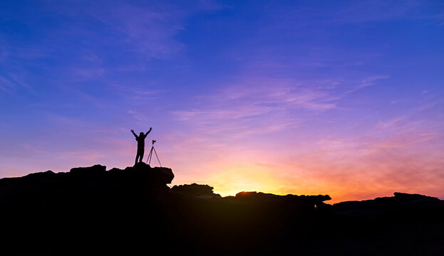 Silhouette of a man photographer standing on top of mountain at sunrise background. The photographer raised hands with the tripod takes photos high in the mountains.