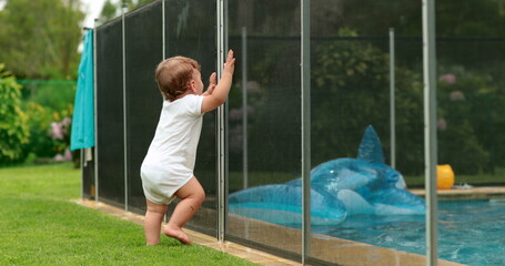Baby leaning on swimming pool fence protection. Infant standing on safety gate