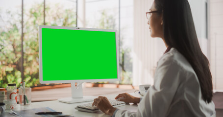 Young Female Doctor Wearing White Coat, Working on Personal Computer with Green Screen Mock Up Display in Her Office. Beautiful Asian Medical Health Care Professional Working with Test Results