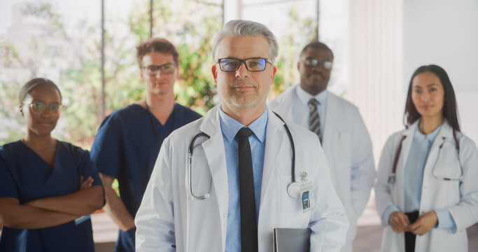 Group of Successful Multiethnic Team of Female and Male Doctors, Nurses and Healthcare Professionals Posing for Camera and Smiling. Portrait of a Confident Middle Aged Physician Standing First in Line