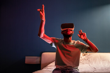 African American man wearing futuristic metaverse VR headset touching air with finger, interacting with virtual objects, shopping in virtual reality while sitting on bed under red neon light