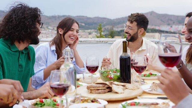 Smiling group of diverse friends having dinner at rooftop party. Happy millennial multiracial people laughing and having fun while drinking and eating sitting on table in summer celebration.