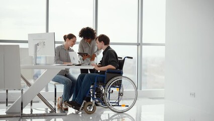 Man with physical disability in wheelchair discuss working issues with colleagues. Teamwork of successful diversity multi ethnic people in modern office. Friendly cooperation, mutual help at work.