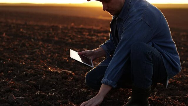 Male farmer touches soil and uses digital tablet for examine and testing cultivated land before sowing on plowed agricultural field on sunset. Smart farming technology and organic agriculture