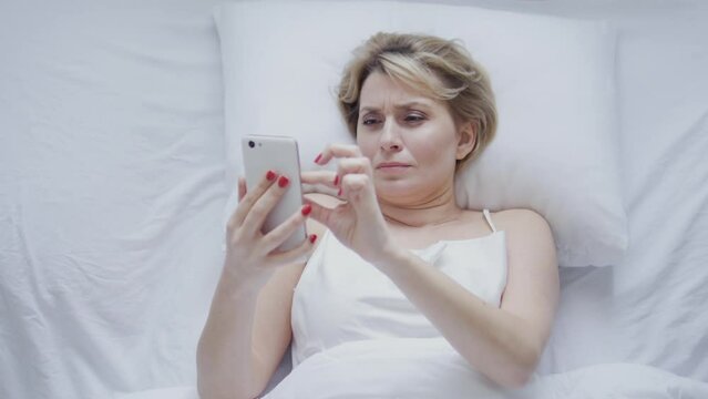 Top view woman scrolling web-pages on smartphone lying in bed late at night, watching posts in social networks and reading news and gossips, evening rituals
