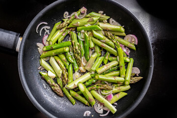 Green asparagus and red onion in a frying pan on the black stove top, cooking a healthy vegetarian meal with seasonal vegetables, high angle view from above