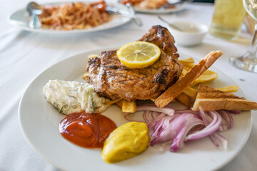 Roasted pork steak with French fries, onions, various dips and a lemon slice, barbecue dish in summer on a white plate, selected focus