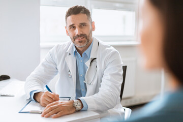 Patient-centered care. Cheerful male doctor consulting lady patient, filling form at consultation in clinic