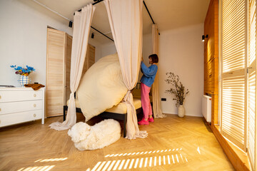 Woman makes a bed while doing some household chores in a cozy bedroom with dog sleeping in front