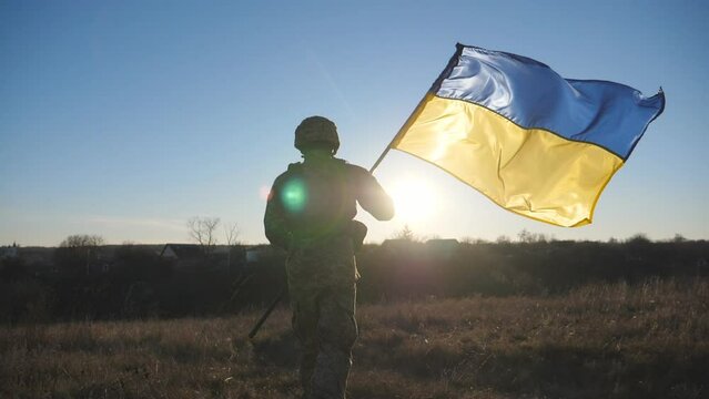 Male soldier of ukrainian army runs with lifted national banner on field. Young man in camouflage uniform jogs with waving flag of Ukraine on meadow at sunset. Invasion resistance concept. Rear view