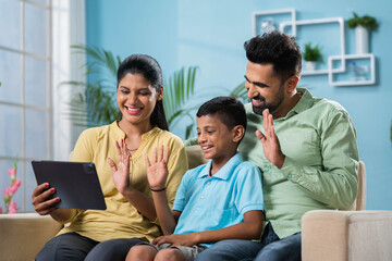 Happy smiling indian couple with kid making video call on digital tablet while sitting on sofa at home - concept of technology, relationship and connection