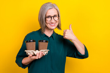 Portrait of attractive lady hold coffee cup order show thumb up wear green isolated on yellow color background