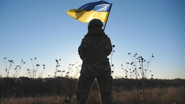 Dolly shot of young girl in military uniform waving flag of Ukraine against background of sunset. Female ukrainian army soldier lifting national banner on the field. Victory against russian aggression